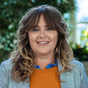 A person with long curly hair smiles at the camera, wearing a grey jacket over an orange top. They are standing in front of a blurred green leafy background.