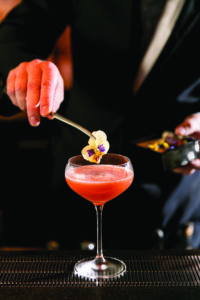 A bartender garnishes a pink cocktail with an edible flower using tweezers. The drink is served in a coupe glass on a bar counter, with a blurred background.