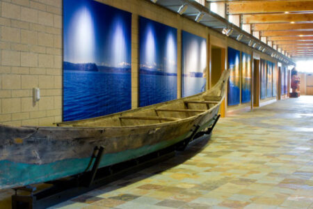 A long, narrow wooden canoe is displayed in a hallway with tiled floors. The wall is adorned with large photographic panels depicting ocean scenes. Exposed wooden beams and brick accent the modern interior.