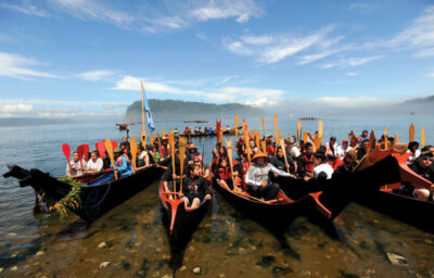 A group of people in several traditional canoes holding paddles, gathered near a shore on a clear day. The sky is blue and the background features a distant island and mist over the water.