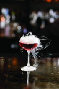 A red cocktail in a coupe glass sits on a wooden bar. White smoke or vapor wafts around the top, creating an atmospheric effect. The background is blurred and dimly lit, enhancing the focus on the drink.