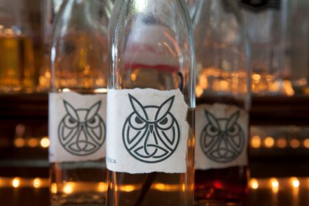 Close-up of glass bottles with labels featuring a stylized owl design. The bottles are arranged in a row with soft, warm lighting in the background, creating a cozy atmosphere.