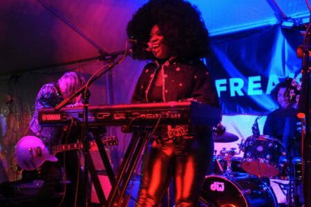 A musician with an afro hairstyle and shiny black outfit plays a keyboard and sings on stage. A guitarist and drummer are in the background under vibrant stage lighting. A banner with partial text 