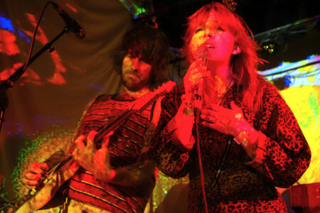 Two musicians perform on stage under colorful, psychedelic lighting. The person on the left plays a guitar while the person on the right sings into a microphone, wearing a patterned outfit. The background features vivid, abstract projections.