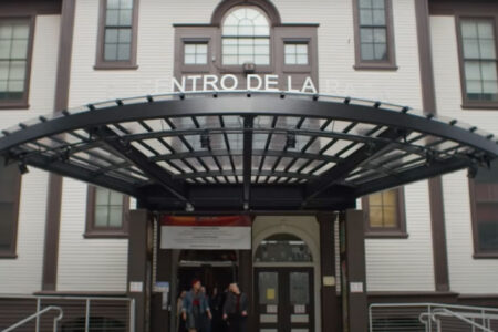 The image shows the entrance to a building with a large, arched glass and metal canopy. The sign above reads "CENTRO DE LA RAZA." Two people are walking out of the building. The exterior is painted in white with dark trim.