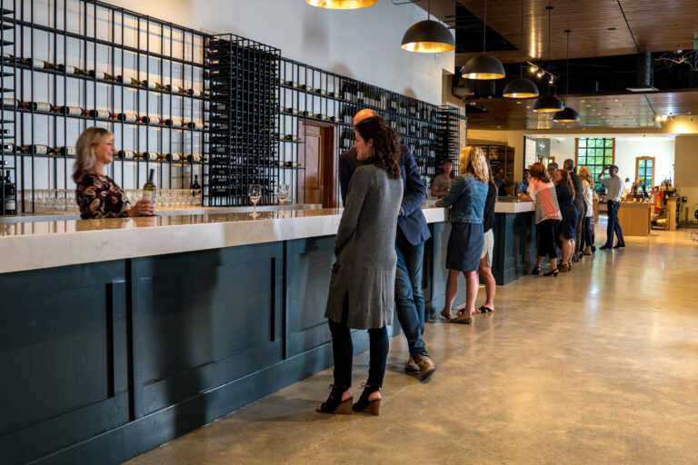 People stand at a long bar counter in a modern Woodinville winery. The bar is adorned with wine bottles in racks. A bartender is serving a customer. The space has a polished concrete floor and pendant lighting, creating a welcoming atmosphere.