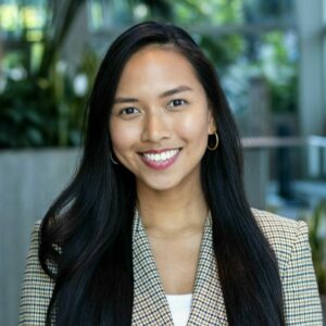 A person with long dark hair and hoop earrings smiles while wearing a checkered blazer, standing indoors with greenery in the background.