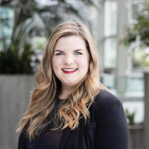 A woman with long blonde hair and a black blazer smiles at the camera. She stands in an indoor setting with blurred greenery and large windows in the background.