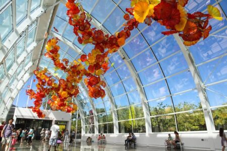 A glasshouse with a high, transparent ceiling houses a vibrant installation of orange, red, and yellow glass flowers. Visitors stroll and sit on benches, enjoying the sunny, lush outdoor view visible through the large windows.