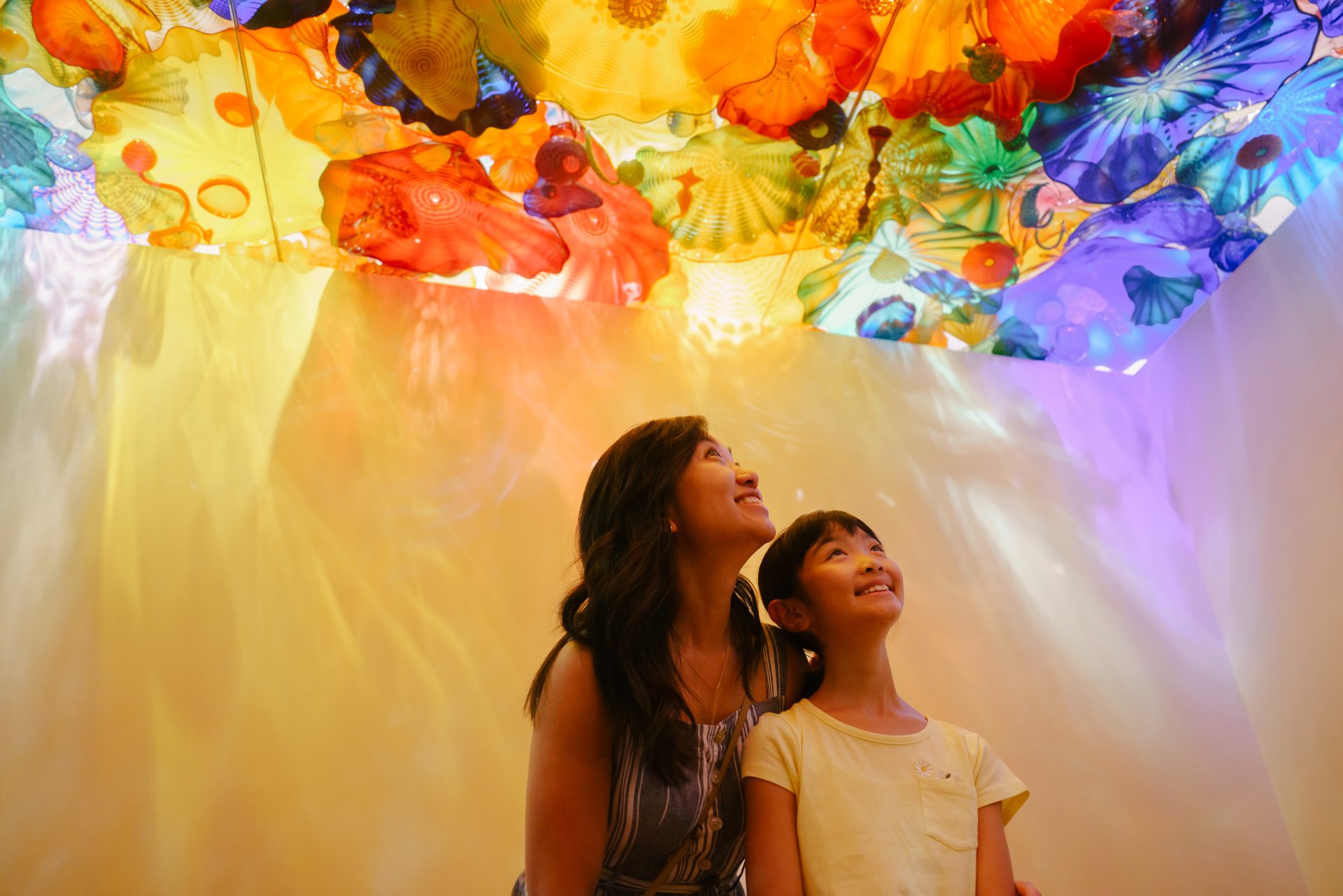 A woman and a young girl gaze up, smiling, at a colorful, translucent ceiling installation resembling glass flowers. The room is bathed in warm, vibrant hues of orange, yellow, blue, and purple.