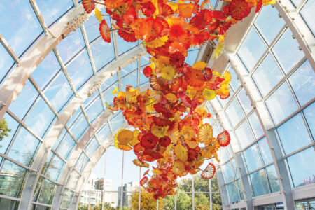 A glass-enclosed atrium with a ceiling adorned with vibrant orange and yellow glass sculptures resembling flowers. People admire the installation, and natural light streams through the tall windows.