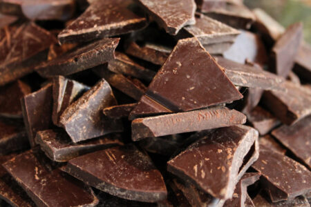 A pile of irregularly shaped dark chocolate pieces, with a rich brown color and slightly rough texture, is shown in close-up. The pieces are stacked on top of each other, highlighting their thickness and varying shapes.