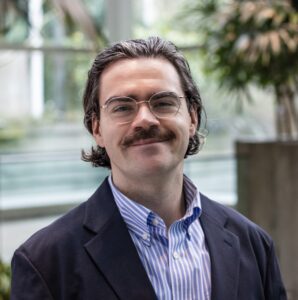 A person with glasses, a mustache, and medium-length hair smiles while wearing a blue striped shirt and dark jacket. They are indoors with a blurred background of plants and large windows.