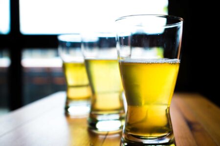 Three half-full glasses of beer sit on a wooden table, lined up in a row. The background shows a blurred window, allowing light to shine through the amber liquid, creating a warm glow.