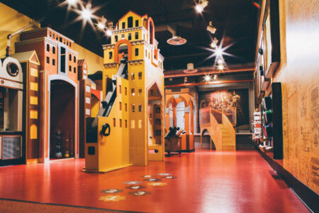 A vibrant indoor play area with colorful, castle-themed structures. The floor is red with scattered game markers, and the walls are decorated with artistic designs. Bright ceiling lights illuminate the playful environment.