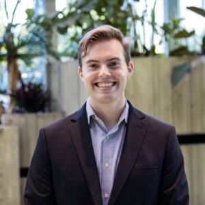 A young man with light brown hair, wearing a dark blazer and a light-colored shirt, smiles broadly while standing indoors in front of large plants and tall windows.