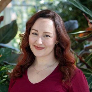 A woman with auburn hair wearing a red top smiles at the camera. She is standing in front of a background filled with lush green plants.