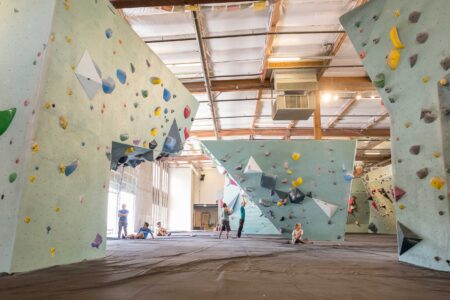 A spacious indoor climbing gym with high walls covered in colorful climbing holds. People are scattered throughout, some climbing and others sitting on the mats. The ceiling features exposed beams and lights, creating an open and industrial feel.