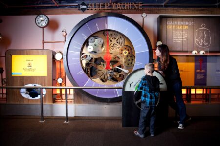 A woman and child interact with a large, colorful display featuring gears and dials, labeled 