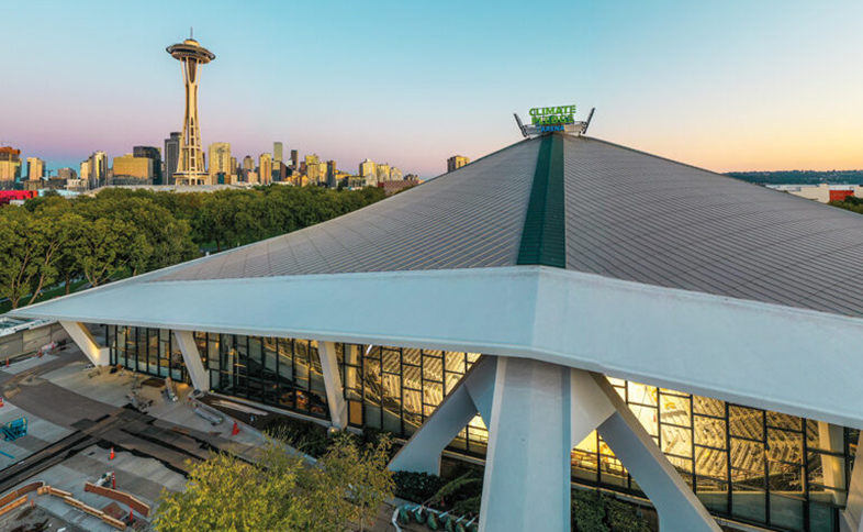Aerial view of the Climate Pledge Arena in Seattle at sunset, featuring its distinctive roof design. The iconic Space Needle and the city's skyline are visible in the background, with trees surrounding the arena.