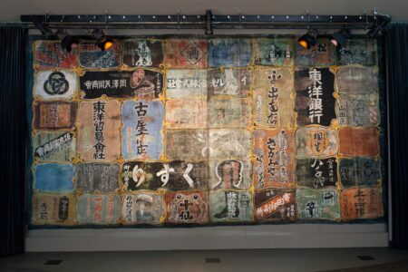 A large, colorful textile wall hanging features numerous vintage Japanese signs and advertisements, displayed in a grid pattern. The signs have diverse designs and kanji characters, set against a gray and blue backdrop.