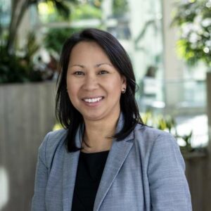 A woman wearing a gray blazer and black top smiles at the camera. She stands in an indoor setting with large potted plants and natural light in the background.