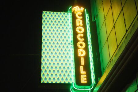 Neon sign for "The Crocodile" glows brightly at night. The sign is vertical, with green and yellow lights, and is mounted on the corner of a building with a patterned facade.