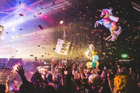 A lively nightclub scene with a crowd dancing under colorful lights. Confetti falls from the ceiling, and there's a large inflatable unicorn and cluster of balloons. People raise their hands, enjoying the vibrant party atmosphere.