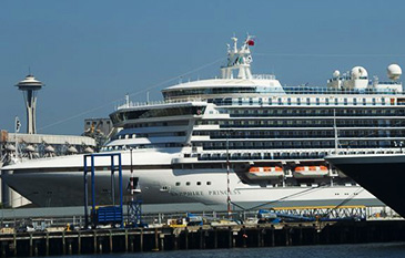 A large cruise ship docked at a port with the Space Needle visible in the background. The ship is white with multiple decks and lifeboats along its side. Clear blue sky above.