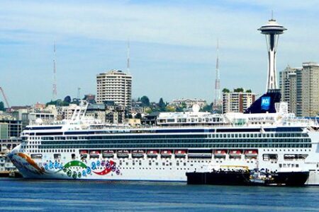 A large cruise ship docked at a port in a city skyline featuring the Space Needle, under a blue sky. Various colorful designs adorn the ship's side. Towering buildings and communication towers are visible in the background.