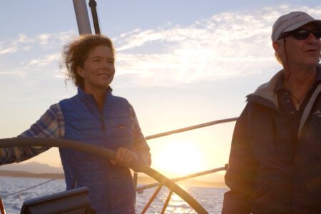 Two people on a sailboat at sunset. One is steering the wheel, wearing a blue vest, while the other, in a dark jacket and cap, stands beside. The sun sets on the horizon, casting a warm glow over the scene.
