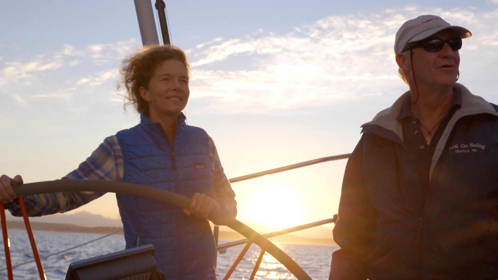 Two people on a sailboat at sunset. One is steering the wheel, wearing a blue vest, while the other, in a dark jacket and cap, stands beside. The sun sets on the horizon, casting a warm glow over the scene.