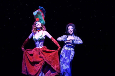 Two performers on stage, one in a vibrant red costume with a feathered headdress, and the other in a blue patterned dress. Both are smiling against a starry background.