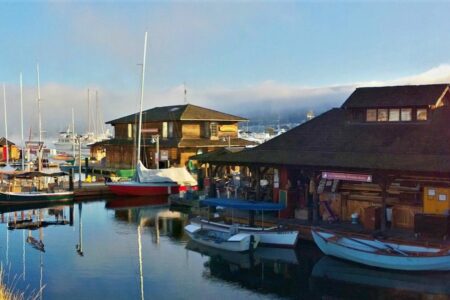 A scenic view of a marina with wooden boats and buildings reflecting on the calm water. The overcast sky adds a misty ambiance to the scene, while the harbor includes a variety of vessels docked alongside rustic structures.