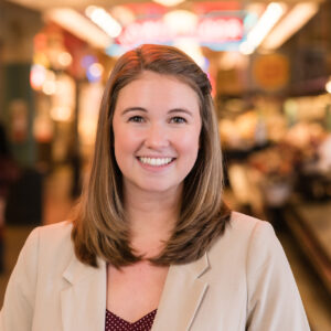 A woman with shoulder-length hair smiles at the camera. She is wearing a beige blazer over a maroon top with white polka dots. The background is a softly focused indoor setting with bright lights and blurred signs.