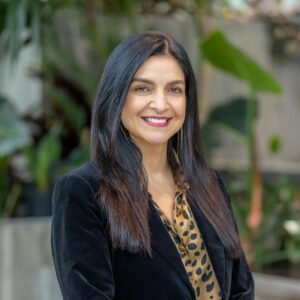 A woman with long dark hair wearing a black blazer and a brown patterned blouse smiles while standing in front of a blurred background with green plants.