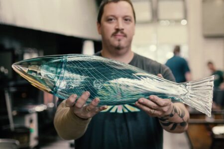 A man holds a large, translucent glass sculpture of a fish in a glassblowing studio. The fish is detailed and intricately designed, with a grid pattern. The background is softly blurred, showing studio equipment and another person.