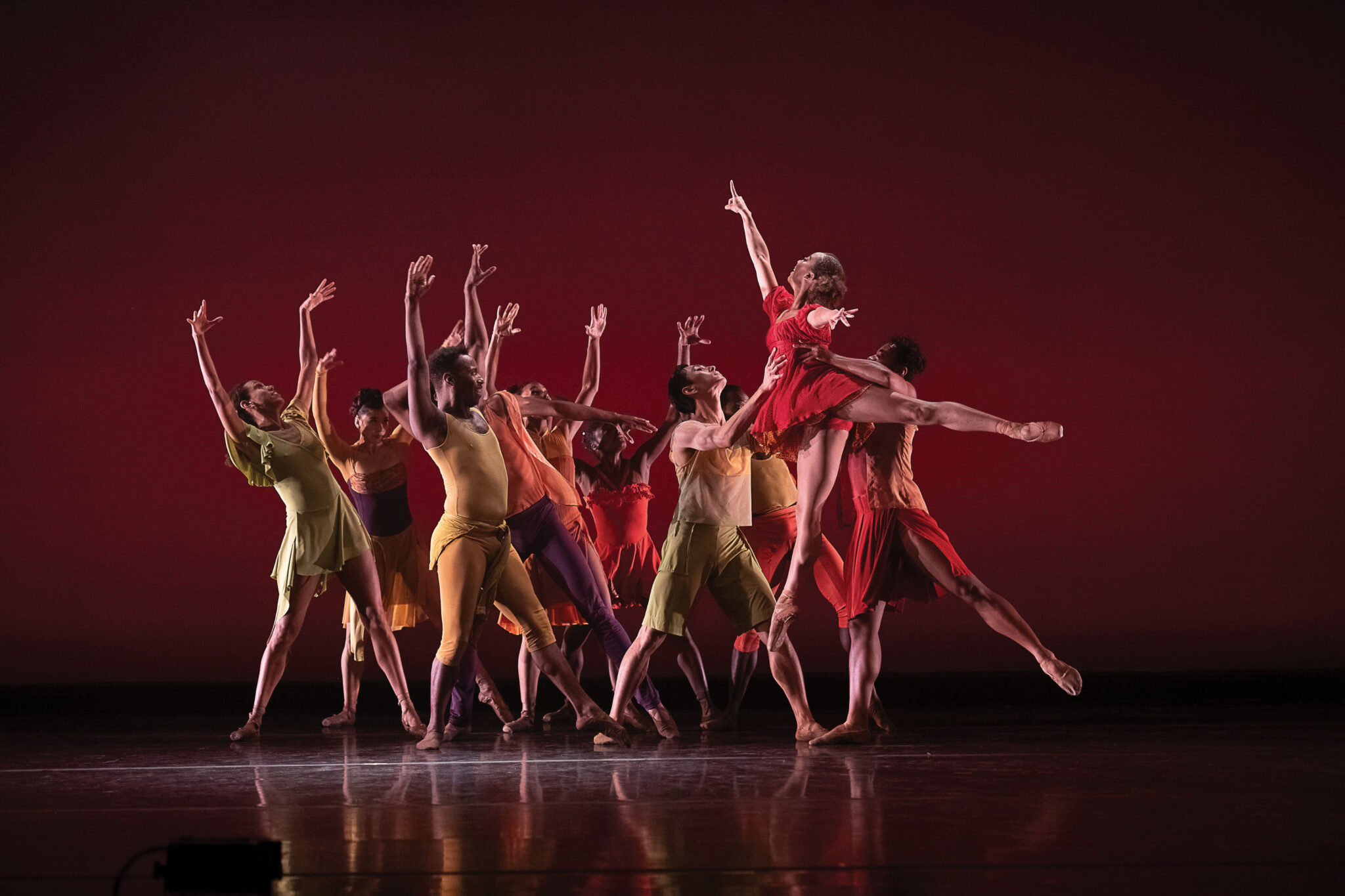 Dancers in colorful outfits perform a dynamic routine on stage. One dancer in red leaps gracefully in the center, while others in yellow, green, and orange strike expressive poses against a deep red backdrop.