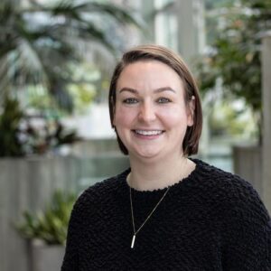 A person with short brown hair and a black knitted sweater smiles at the camera. They are wearing a necklace and standing in a bright room with green plants in the background.