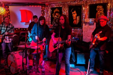 A band performs on stage at a dimly lit venue. Five musicians play instruments including guitars and drums. A backdrop of silver tinsel and framed portraits adds color. A screen displays the letter 