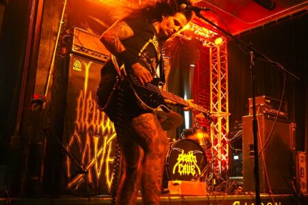 A guitarist with long hair and tattoos energetically plays on stage at a venue called "Central Saloon." The stage is lit with vibrant orange and red lights, and a drum set and amplifiers are visible in the background.