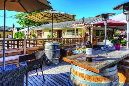 A sunny outdoor patio featuring barrel tables with wine glasses and a bottle. Wicker chairs surround the tables, and large umbrellas provide shade. A wooden fence borders the area, and more seating is visible in the background.