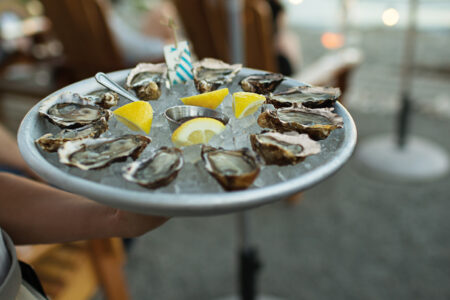 A person holds a platter of fresh oysters on ice with lemon wedges and a small dipping sauce by the waterfront. Wooden chairs and a view of boats in the harbor are in the background.
