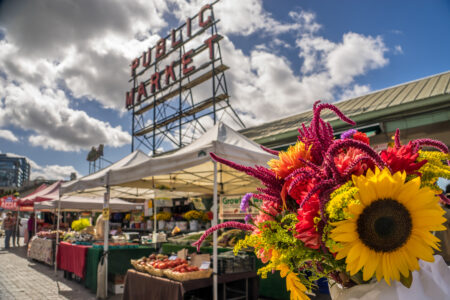 A vibrant bouquet of sunflowers and other colorful flowers in the foreground with a bustling outdoor market scene behind. The iconic "Public Market" sign is visible, under a partly cloudy sky.