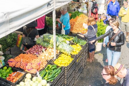 Colorful produce fills stands under white tents with people walking by at a a farmers market.