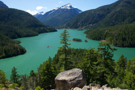 A stunning view of a turquoise lake surrounded by lush green forests and towering mountains, covered in patches of snow. A rocky foreground overlooks the serene landscape under a clear blue sky.