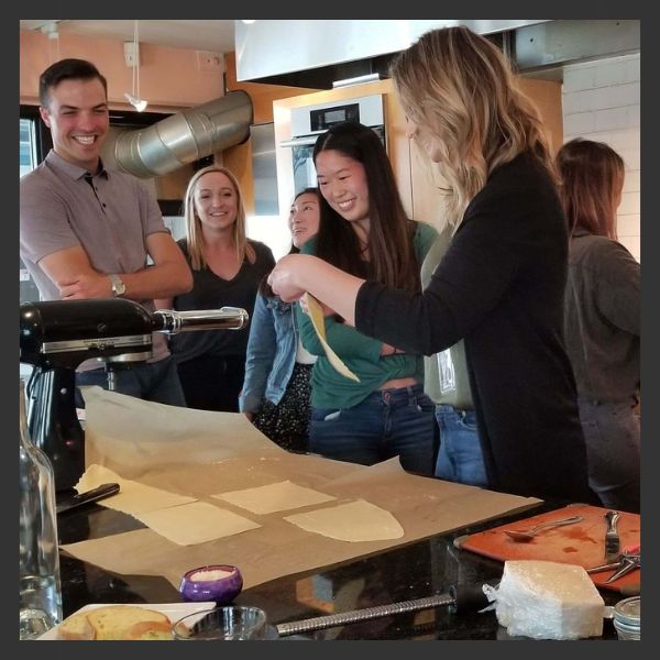 A group of people in a kitchen smile and watch as one woman feeds sheets of dough through a pasta maker on a countertop covered with parchment and cooking tools.