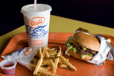 A tray with a cheeseburger wrapped in foil, a side of fries, a small cup of ketchup, and a large milkshake in a cup labeled "Dick's.