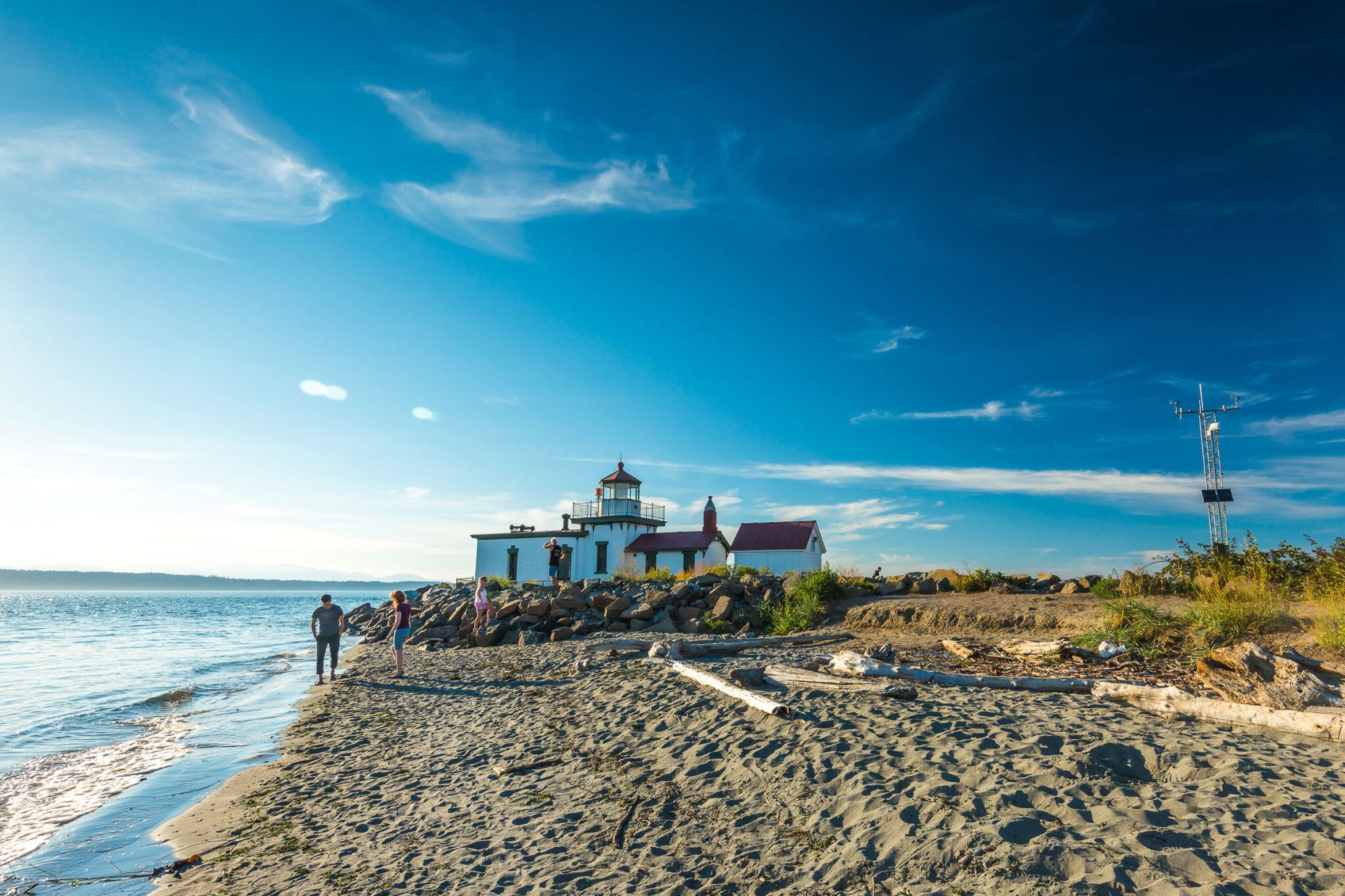 A picturesque coastal scene with a lighthouse surrounded by rocky terrain and grass. A clear blue sky and ocean enhance the view. People walk along the sandy beach, creating a serene, sunny atmosphere.