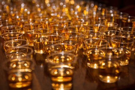 Rows of small glasses filled with amber-colored liquid are arranged on a wooden table. The glasses reflect warm, golden hues, creating a glowing effect. The background is softly blurred, emphasizing the drinks in the foreground.
